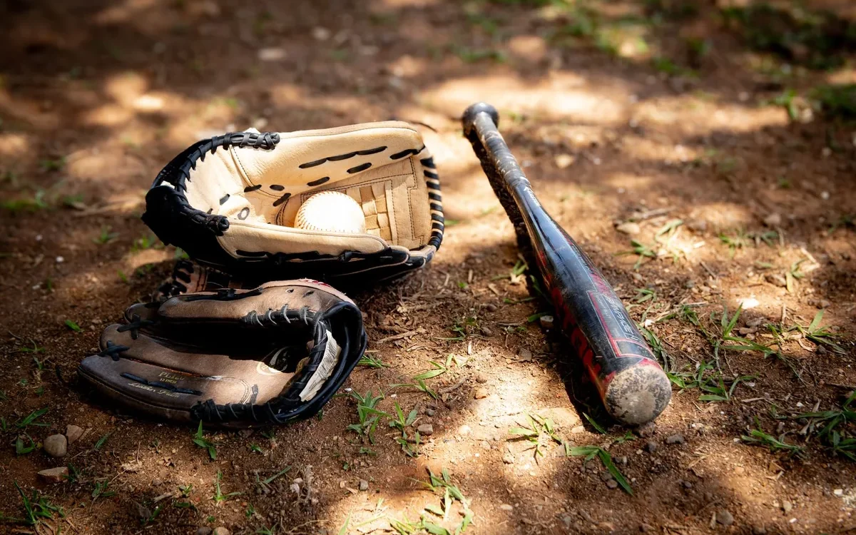 Două mănuși de baseball, o minge albă și o bâtă neagră așezate pe pământ