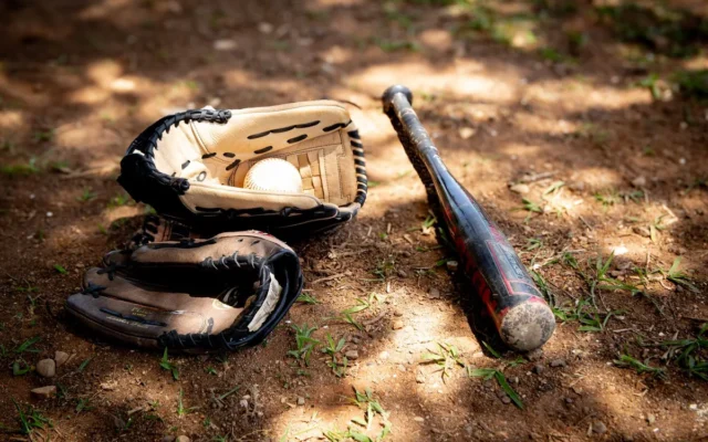 Două mănuși de baseball, o minge albă și o bâtă neagră așezate pe pământ