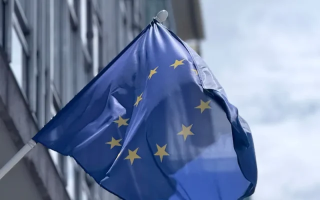 Drapel albastru al Uniunii Europene cu stele galbene în fața unei clădiri cu geamuri