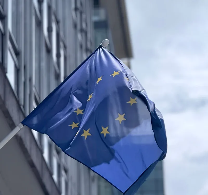 Drapel albastru al Uniunii Europene cu stele galbene în fața unei clădiri cu geamuri