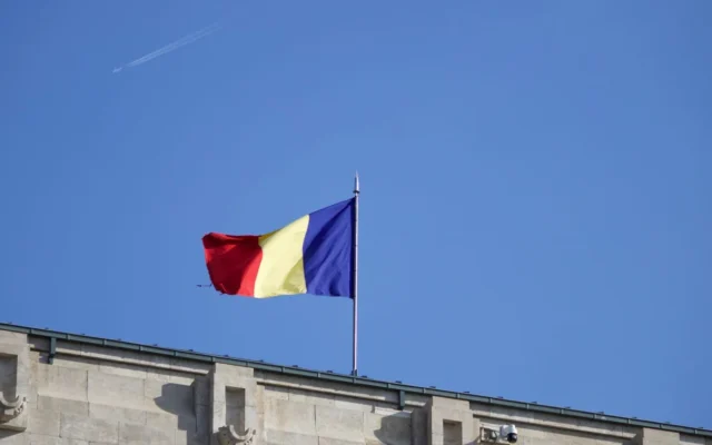 Drapelul României pe un catarg deasupra unei clădiri, pe fundal de cer albastru