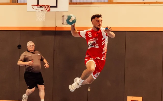 Jucător de handbal în echipament roșu și alb, în săritură cu mingea într-o sală de sport