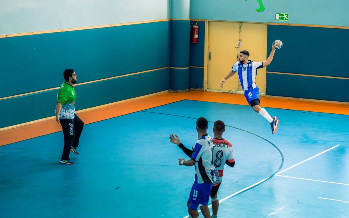 Jucător de handbal în săritură cu mingea, în fața porții și a altor jucători, pe un teren albastru într-o sală