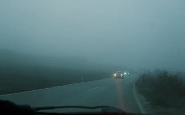 Mașini cu farurile aprinse pe un drum în curbă, acoperit de ceață