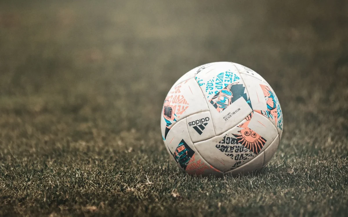 Minge de fotbal albă, cu desene portocalii, turcoaz și negre, așezată pe gazon
