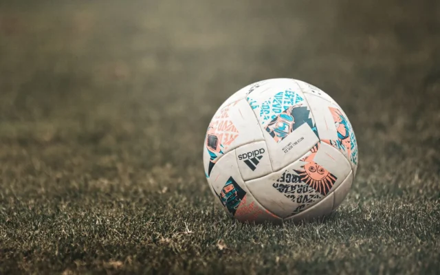 Minge de fotbal albă, cu desene portocalii, turcoaz și negre, așezată pe gazon