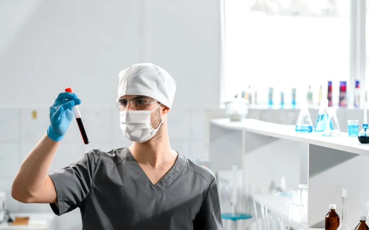 Persoană cu echipament de protecție ținând o eprubetă cu sânge într-un laborator