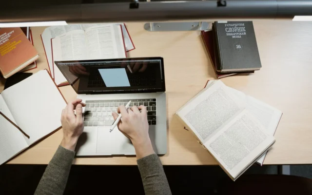 Mâinile unei persoane la un laptop, cu cărți și caiete pe birou.