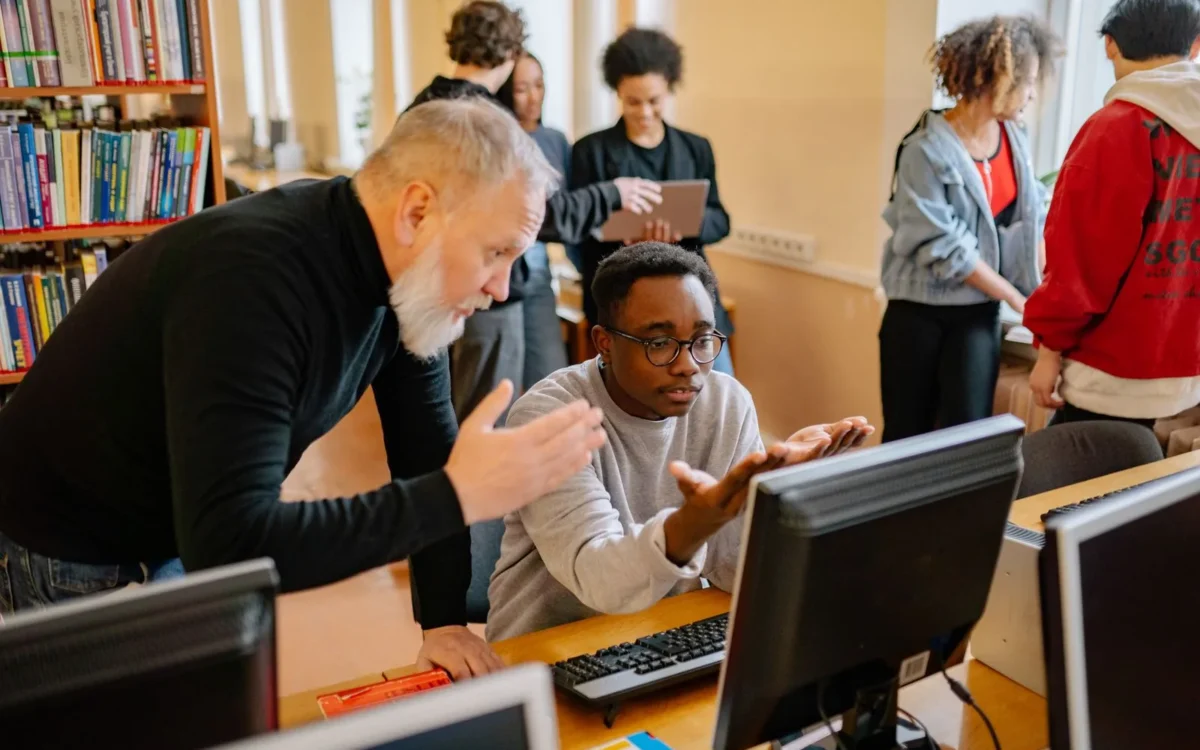 Profesor și cursant în fața unui computer, lângă rafturi cu cărți