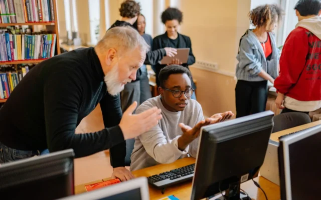 Profesor și cursant în fața unui computer, lângă rafturi cu cărți