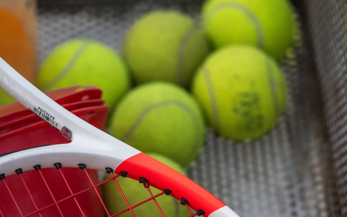 Rachetă de tenis albă și roșie lângă un coș metalic cu mingi galbene
