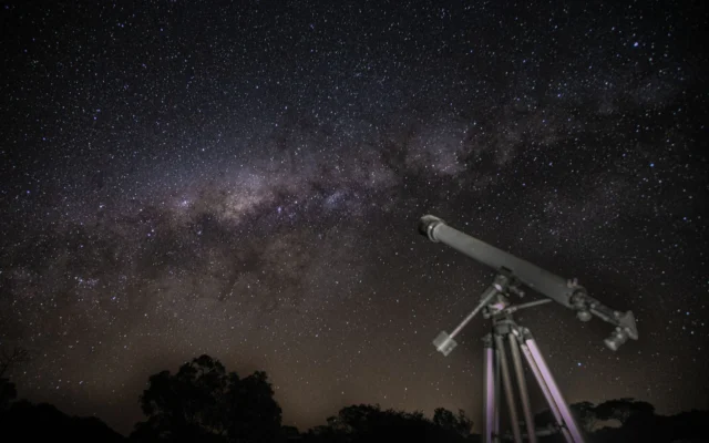 Telescop pe trepied sub un cer înstelat, cu siluete de arbori la orizont