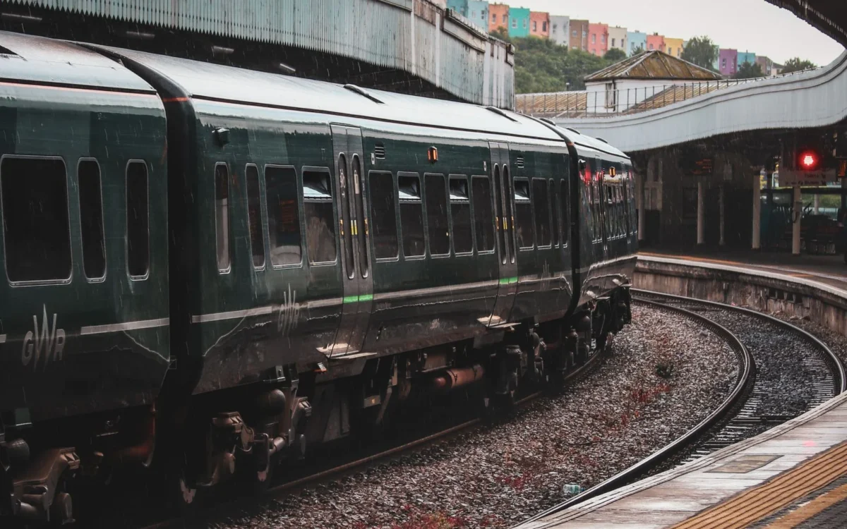 Tren verde pe șine curbate lângă un peron acoperit, cu clădiri în fundal