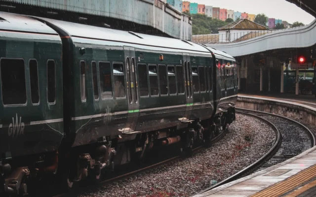 Tren verde pe șine curbate lângă un peron acoperit, cu clădiri în fundal