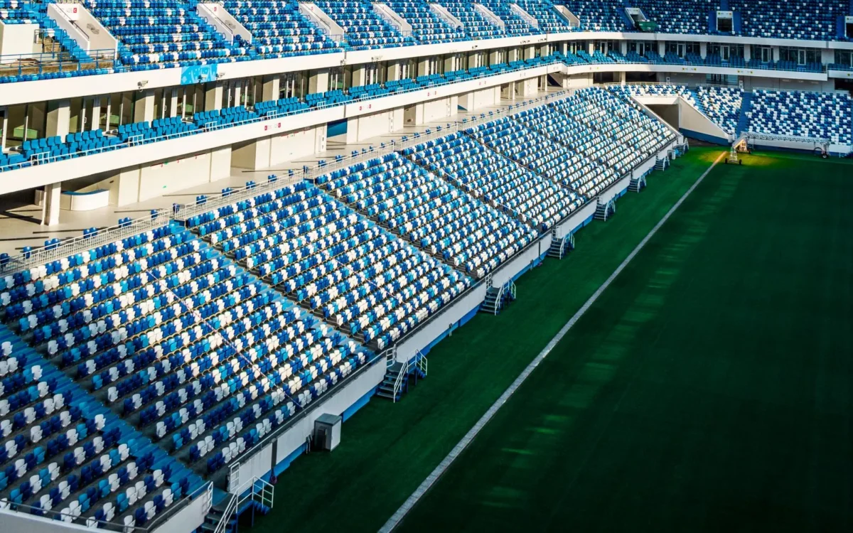 Tribune goale cu scaune albastre și albe și gazonul unui stadion de fotbal