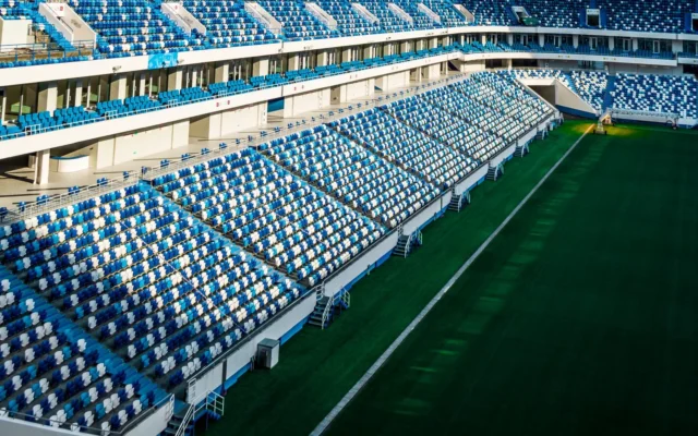 Tribune goale cu scaune albastre și albe și gazonul unui stadion de fotbal